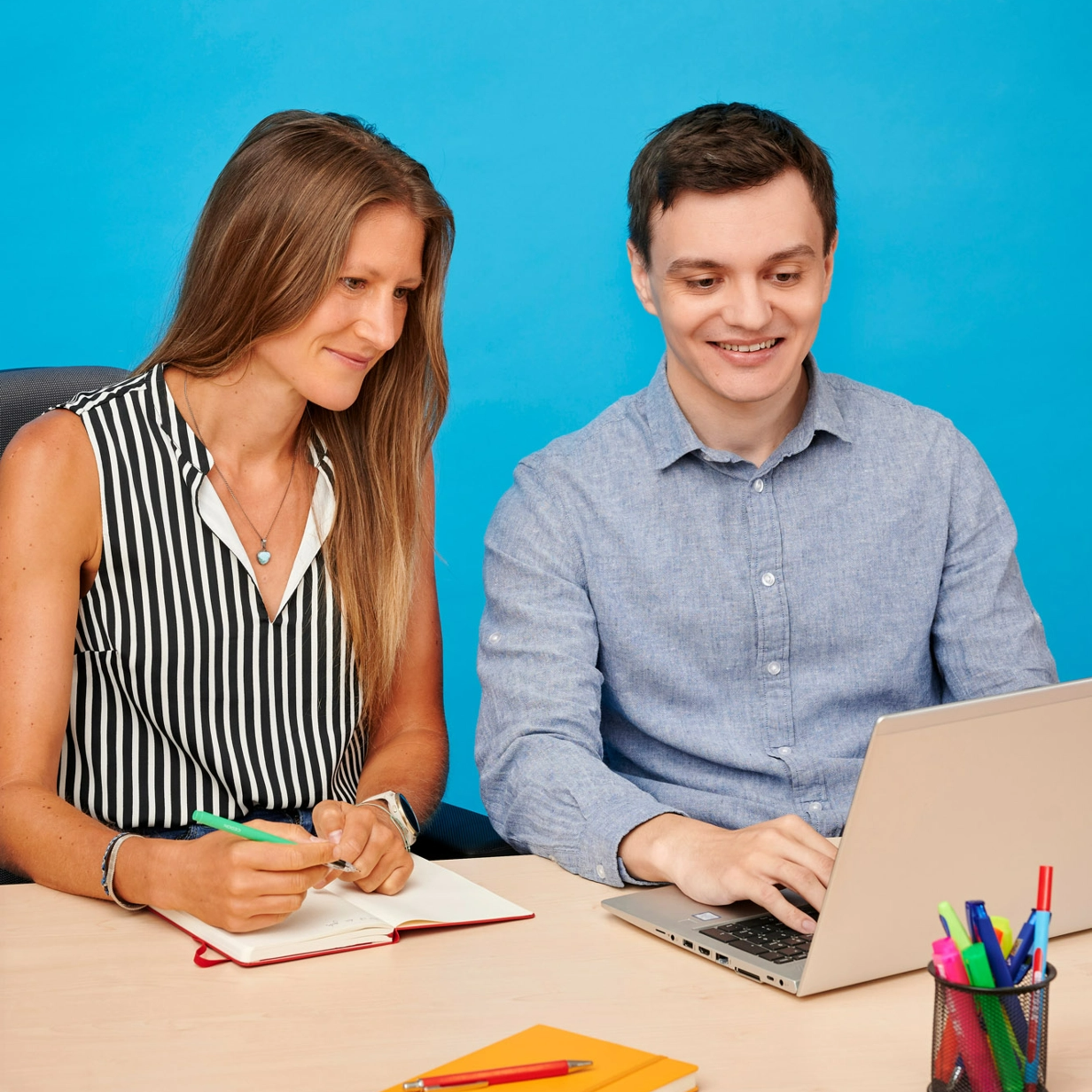 Das Bild zeigt zwei Personen, einen jüngeren Mann und eine mittelalte Frau. Der junge Mann sitzt am Schreibtisch und arbeitet freundlich schauend an einem Laptop. Die Frau sitzt links neben ihm und schaut ihm zu und lässt sich etwas erklären. Die Frau hat einen Stift in der Hand und ein Notizbuch vor sich. Die Szenerie ist bunt. Es liegen bunte Accessoires auf dem Tisch. Der Hintergrund ist eine blaue Wand.