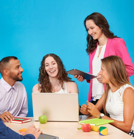 Das Bild zeigt vier Personen zwischen 20 und 30 Jahren. Drei von ihnen sitzen an einem Tisch um ein Laptop herum. Eine Person steht neben dem Tisch. Alle arbeiten bei der FinanzIT BAYERN und reden und lachen. Die Szenerie ist bunt. Es liegen bunte Accessoires auf dem Tisch. Der Hintergrund ist eine blaue Wand.