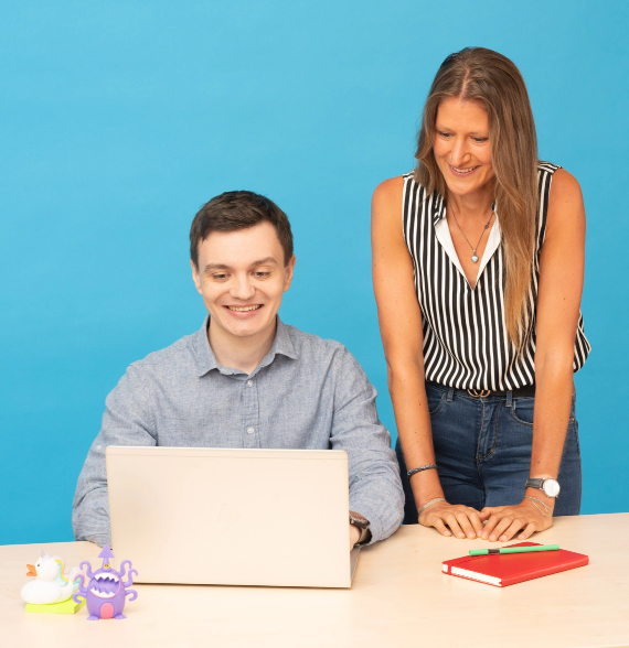 Das Bild zeigt zwei Personen, einen jüngeren Mann und eine mittelalte Frau. Der junge Mann sitzt am Schreibtisch und arbeitet freundlich schauend an einem Laptop. Die mittelalte Frau steht neben ihm und erklärt ihm etwas oder lässt sich etwas erklären. Beide Personen arbeiten bei der FinanzIT BAYERN und lachen. Die Szenerie ist bunt. Es liegen bunte Accessoires auf dem Tisch. Der Hintergrund ist eine blaue Wand