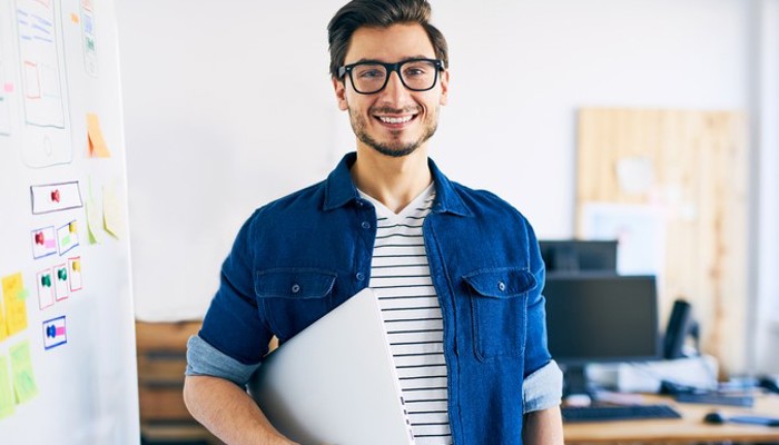 Bild zeigt lächelnden jungen Mann mit Brille und Laptop unter dem rechten Arm